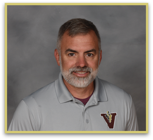 A person with short hair and a beard is smiling and wearing a light gray polo shirt with a maroon and gold “V" logo on the chest against a gray background. Description generated by AI