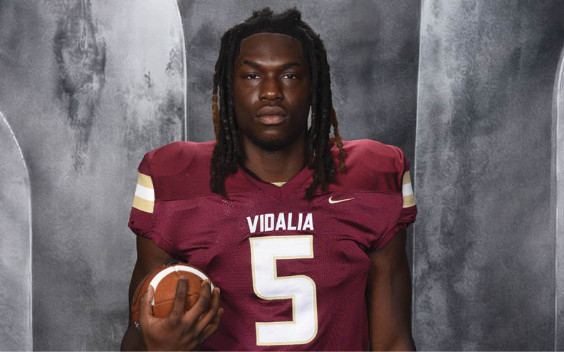 A person wearing a maroon football jersey with the number five and the word Vidalia is holding a football against a gray backdrop. Description generated by AI