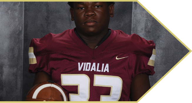 A person wearing a maroon football jersey with the number 33 and the word Vidalia is holding a football against a dark background. Description generated by AI
