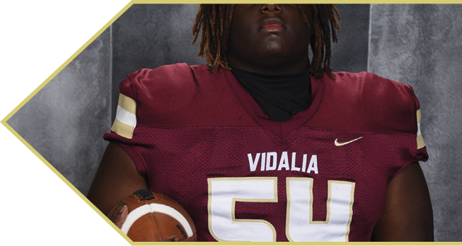 A person wearing a maroon football jersey with the name “VIDALIA" and number 54 is holding a football against a gray background. Description generated by AI