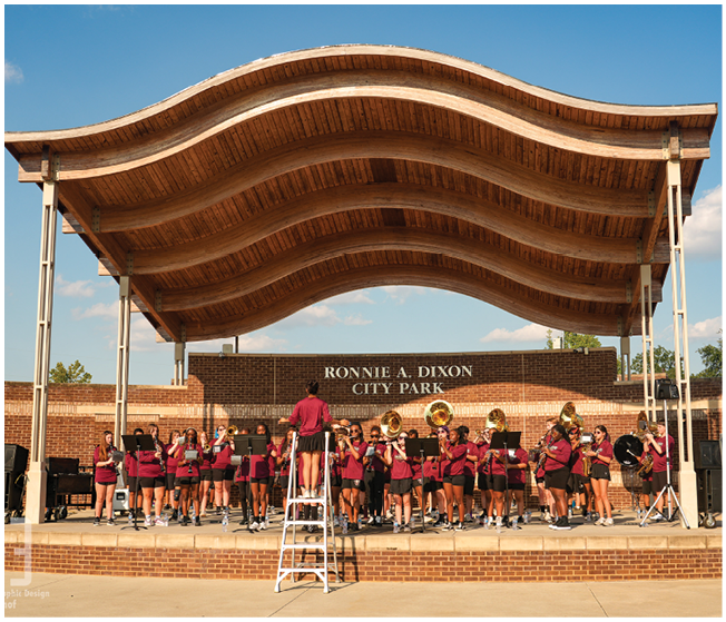 A group of people wearing matching maroon shirts perform music with brass and woodwind instruments under a wooden wave-shaped pavilion at Ronnie A. Dixon City Park. Description generated by AI
