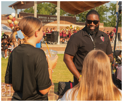 Three adults are engaged in conversation outdoors near a stage with a band playing and balloons in the background at a park event. Description generated by AI