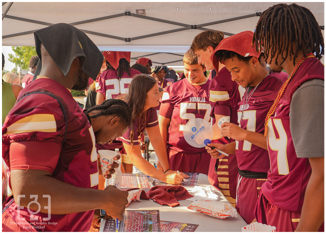 People wearing matching sports jerseys gather around a table under a canopy, engaging in signing or organizing items during an outdoor event. Description generated by AI