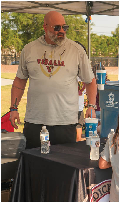 An adult wearing sunglasses and a light-colored shirt with “VIDALIA" printed on it stands behind a table with water bottles and clear cups under a canopy outdoors. Description generated by AI