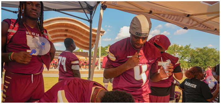 People wearing matching sports jerseys are gathered under a tent at an outdoor event with a stadium and trees visible in the background. Description generated by AI