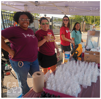 Four people stand behind a table with plastic bags and cleaning supplies outdoors, smiling and wearing casual clothing and sunglasses. Description generated by AI