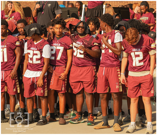 A group of people wearing matching maroon sports uniforms stand together outdoors in front of a brick wall, some looking at each other and others looking down. Description generated by AI