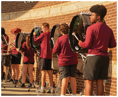 A group of people wearing matching maroon shirts and black shorts are playing various percussion instruments outdoors against a brick wall. Description generated by AI