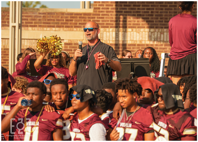 A group of people wearing matching sports jerseys and a person holding a microphone stand together outdoors near a brick wall, with one person holding gold pom-poms. Description generated by AI
