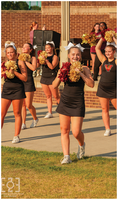 People wearing black outfits with white bows are holding gold and red pom-poms and appear to be performing a cheer routine outdoors near a brick wall and large speakers. Description generated by AI