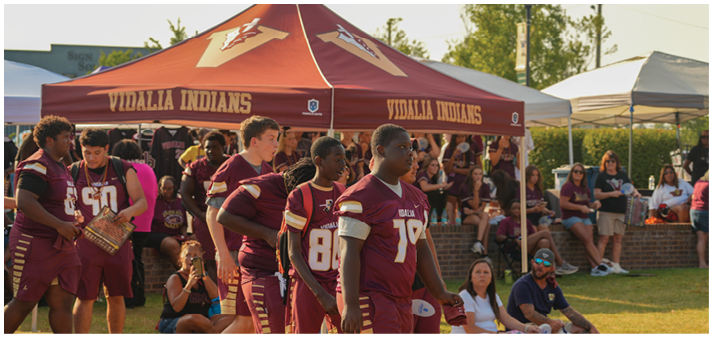 People wearing maroon and gold football uniforms stand near a tent labeled “Vidalia Indians" with spectators seated in the background. Description generated by AI
