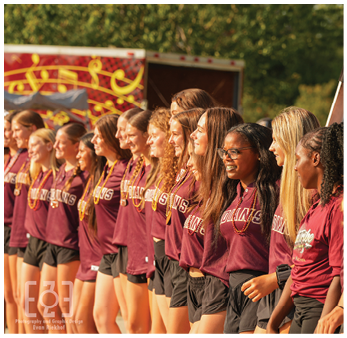 A line of people wearing matching maroon shirts and black shorts stand side by side outdoors, smiling and facing the same direction. Description generated by AI