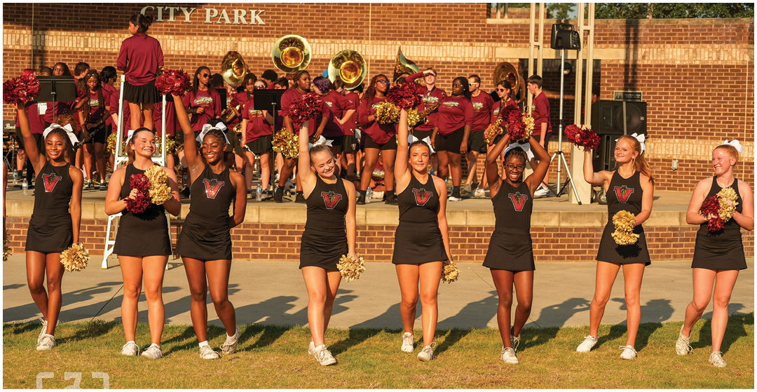 A group of adults in matching cheerleading uniforms hold pom-poms and smile while standing on grass in front of a marching band performing on a brick stage. Description generated by AI