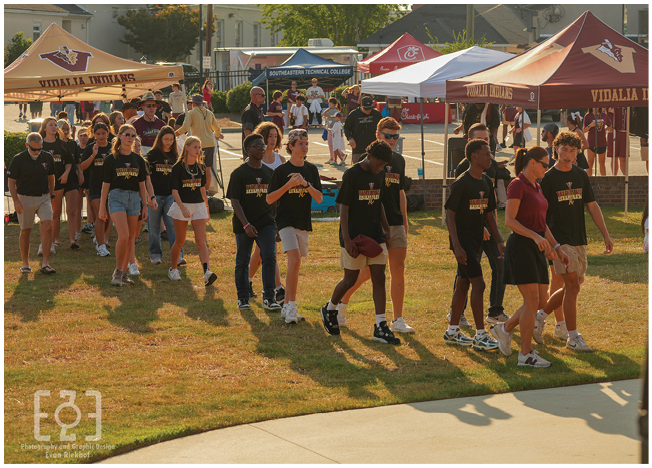 A group of people wearing matching black shirts and casual shorts or pants are walking on grass near tents with logos and text in an outdoor event setting. Description generated by AI