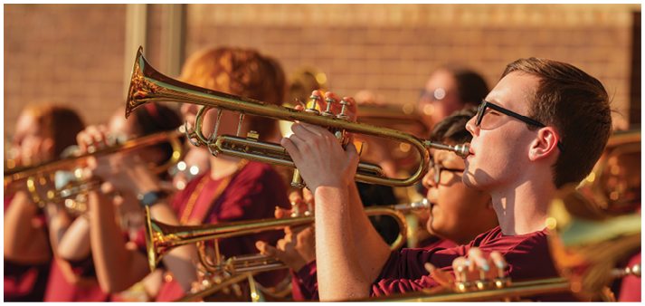 A group of adults wearing matching maroon shirts are playing trumpets outdoors in a coordinated formation. Description generated by AI