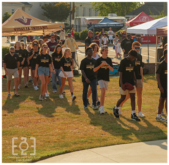 A group of people wearing matching black shirts walk together on grass near tents with logos and banners in an outdoor event setting. Description generated by AI