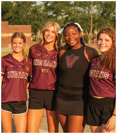 Four smiling people wearing sports uniforms stand closely together outdoors with trees and a brick wall in the background. Description generated by AI