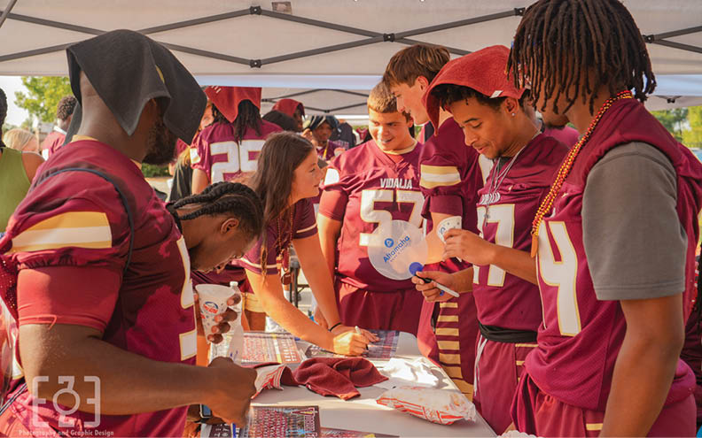 People wearing sports jerseys gather under a tent, engaging in conversation and signing items on a table. Description generated by AI