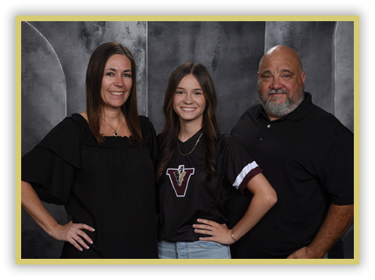 Three people stand closely together smiling against a dark textured backdrop, with the person in the middle wearing a black shirt with a large “V" logo and light blue jeans, while the others wear black tops. Description generated by AI