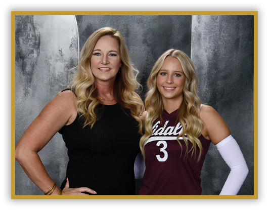 Two adults with long wavy hair stand side by side smiling, one wearing a black sleeveless dress and the other in a maroon sports jersey with the number three and a white arm sleeve, against a gray textured backdrop. Description generated by AI