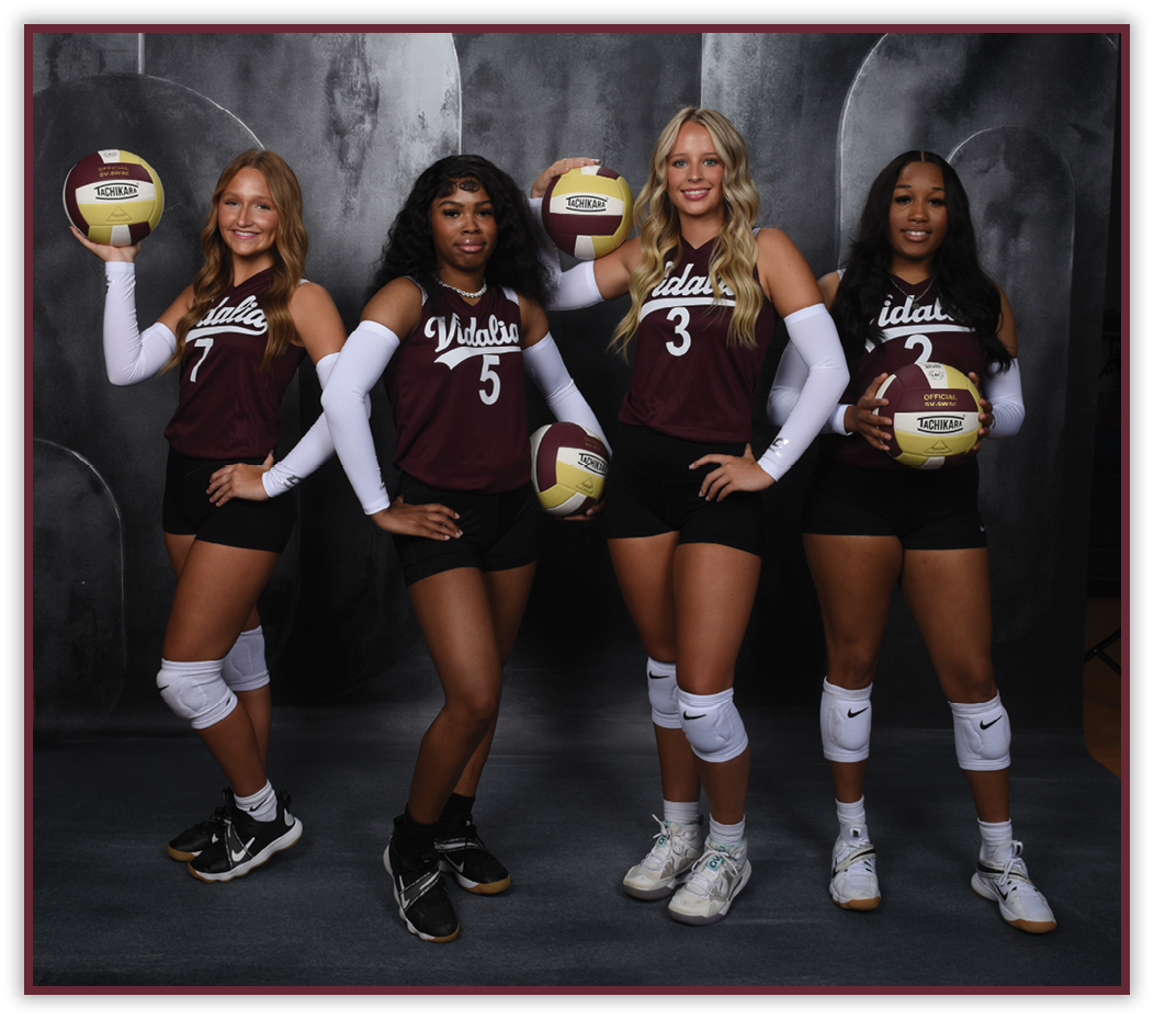 Four adults wearing matching sports uniforms and knee pads pose holding volleyballs against a dark backdrop. Description generated by AI