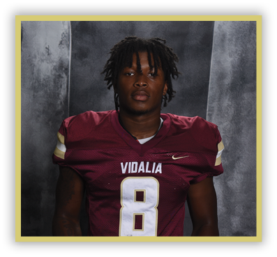 A person with short dreadlocks wearing a maroon football jersey with the word Vidalia and the number 8 stands against a dark gray backdrop. Description generated by AI