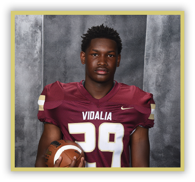 A person wearing a maroon football jersey with the number 29 and the word Vidalia is holding a football against a gray backdrop. Description generated by AI