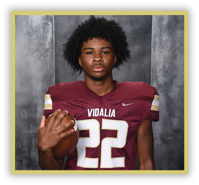 Person with curly hair wearing a maroon football jersey with the number 22 and holding a football against a gray backdrop. Description generated by AI