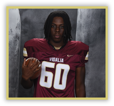 A person with shoulder-length dreadlocks wearing a maroon football jersey with the number 60 and the word Vidalia is holding a football against a gray backdrop. Description generated by AI
