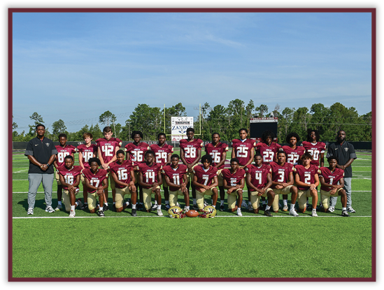 A group of people wearing maroon and gold football uniforms pose for a team photo on a green football field with two coaches standing on either side. Description generated by AI