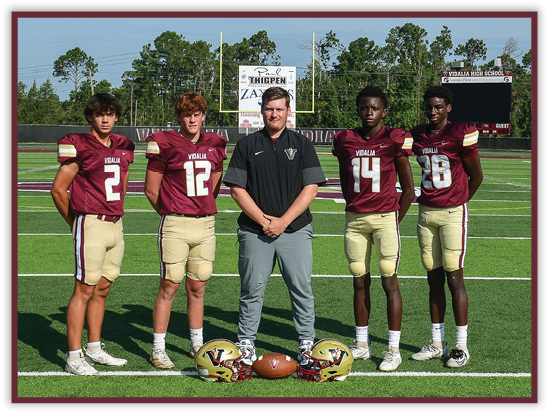Five people wearing football uniforms and one person in casual sportswear stand on a football field with helmets and a football placed on the ground in front of them. Description generated by AI