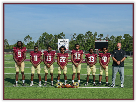 Seven people wearing matching football uniforms stand in a line on a football field with helmets and a football placed on the ground in front of them and a scoreboard visible in the background. Description generated by AI