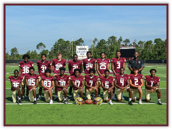 A group of people wearing maroon and gold football uniforms pose together on a football field with trees and a scoreboard in the background. Description generated by AI