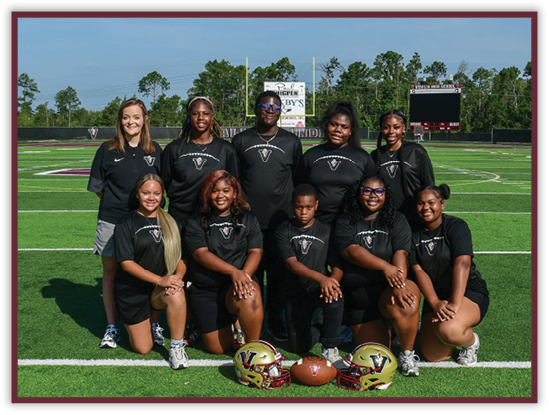 A group of adults and one child wearing matching black sports shirts pose on a football field with two helmets and a football placed in front of them. Description generated by AI