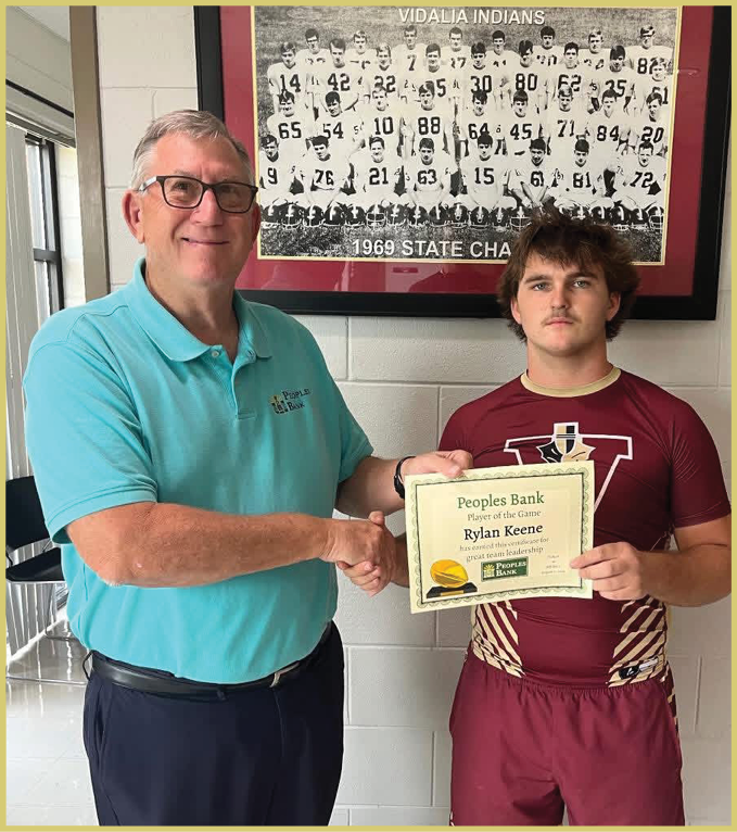 Two people stand in front of a framed black and white team photo labeled “Vidalia Indians 1969 State Champs" on a wall. Description generated by AI