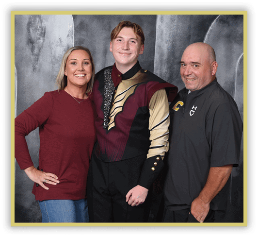 Three adults stand closely together smiling, with one wearing a maroon and gold uniform and the others dressed casually against a gray backdrop. Description generated by AI