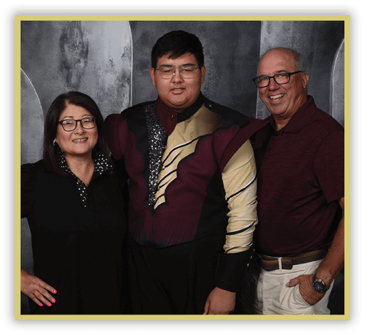 Three adults stand closely together smiling against a dark textured backdrop, with the person in the center wearing a maroon and gold uniform. Description generated by AI
