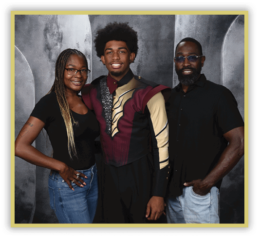 Three adults stand closely together smiling, with one wearing a decorative maroon and gold uniform, and the others dressed casually against a gray textured backdrop. Description generated by AI
