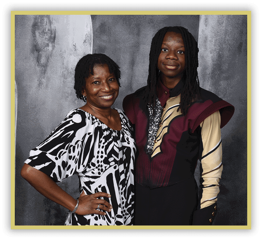 Two people stand closely together smiling, one wearing a black and white patterned dress and the other wearing a maroon and beige costume with decorative details. Description generated by AI