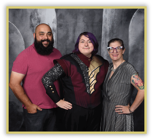 Three people stand closely together smiling, with one wearing a maroon and black costume with decorative details, another in a pink shirt, and the third in a striped dress with colorful tattoos on one arm and large glasses. Description generated by AI
