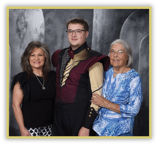 Three people stand closely together against a gray backdrop, with one person in the center wearing a maroon and black uniform with gold accents, while the others wear casual clothing and smile. Description generated by AI
