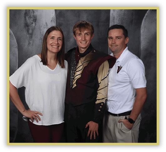 Three adults stand closely together smiling, with one wearing a marching band uniform and the others dressed in casual and semi-casual clothing against a dark backdrop. Description generated by AI