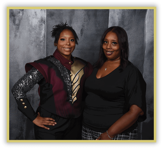 Two adults stand side by side against a dark textured background, one wearing a decorative costume with sequins and metallic accents and the other dressed in a black top and patterned pants. Description generated by AI