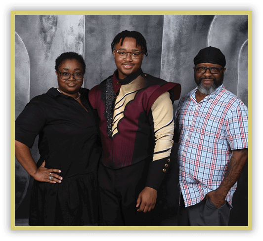 Three adults stand closely together smiling, with one wearing a maroon and beige uniform, another in a black dress, and the third in a plaid shirt and head covering against a gray textured backdrop. Description generated by AI