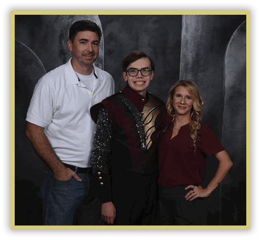 Three people stand closely together smiling, with one person in the center wearing a decorative uniform and the other two dressed casually against a dark backdrop. Description generated by AI