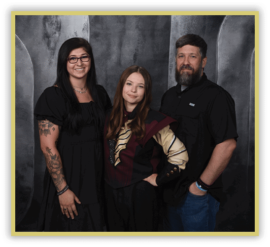 Three adults stand closely together against a dark textured backdrop, with one wearing glasses and a black dress showing tattoos, another in a maroon and beige patterned top, and the third in a black shirt and jeans smiling. Description generated by AI