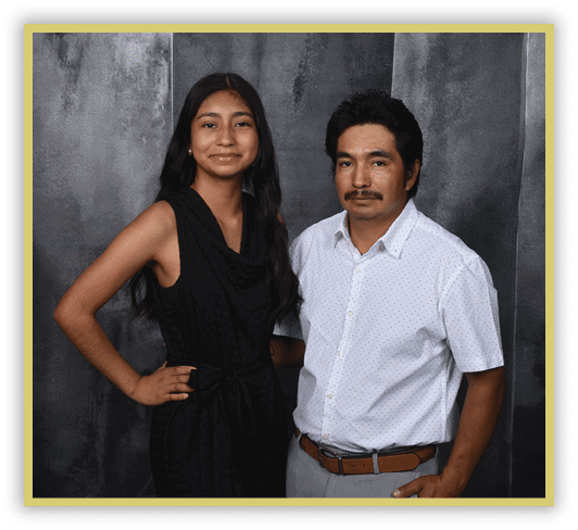 Two adults stand side by side against a dark textured background, one wearing a sleeveless black outfit and the other wearing a white short-sleeve button-up shirt with light-colored pants and a brown belt. Description generated by AI