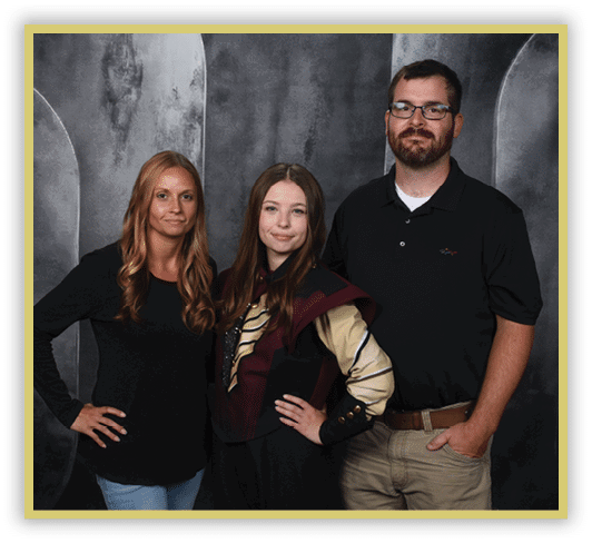 Three adults stand closely together against a dark textured backdrop, with one wearing a black shirt and glasses, another in a black long-sleeve top and light jeans, and the third dressed in a costume with a maroon and beige jacket. Description generated by AI
