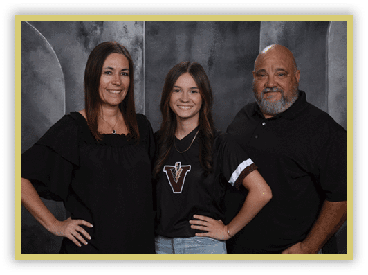 Three people stand closely together smiling against a dark textured backdrop, with the person in the middle wearing a black shirt with a large “V" logo and light blue jeans, while the others wear black tops. Description generated by AI