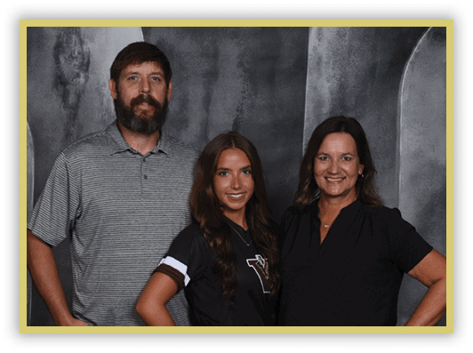 Three adults stand closely together smiling against a dark textured backdrop, with one wearing a striped polo shirt, another in a black sports jersey, and the third in a black blouse. Description generated by AI